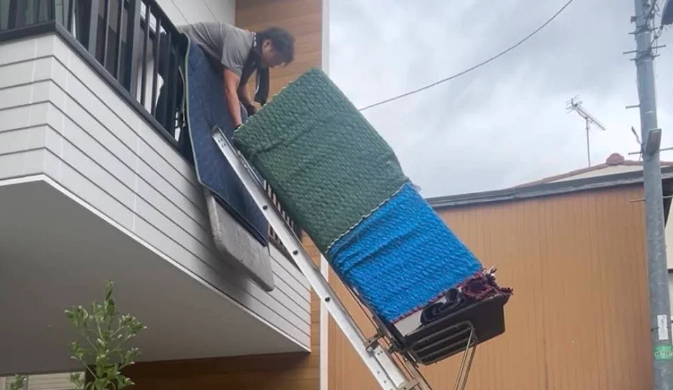 Workers carrying a large bedding item out from a balcony using a powered ladder along the exterior wall