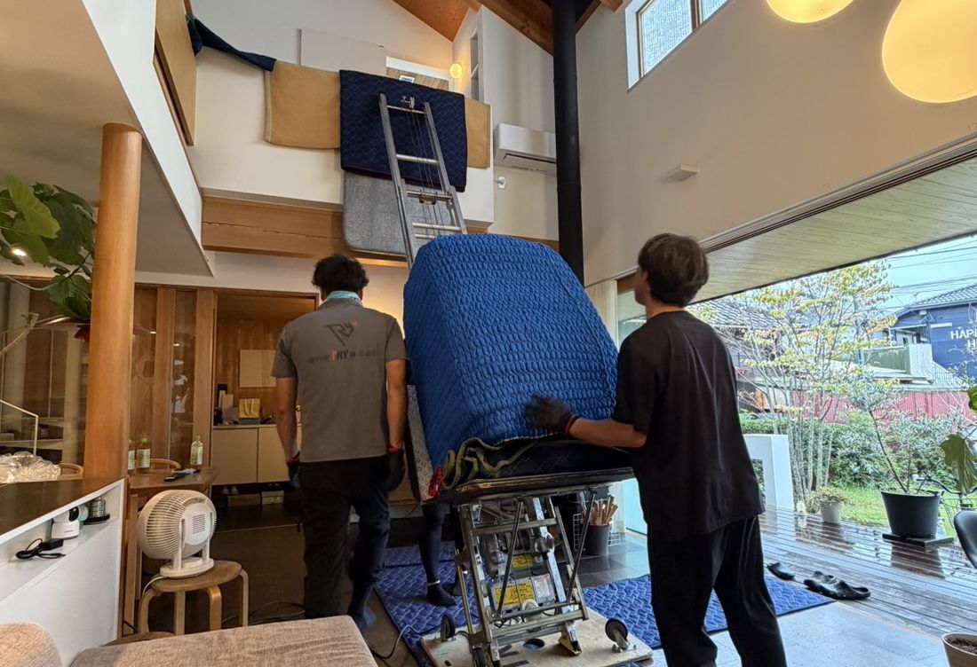 Two workers carefully carrying a large piece of furniture protected with blue padding through an open-plan interior