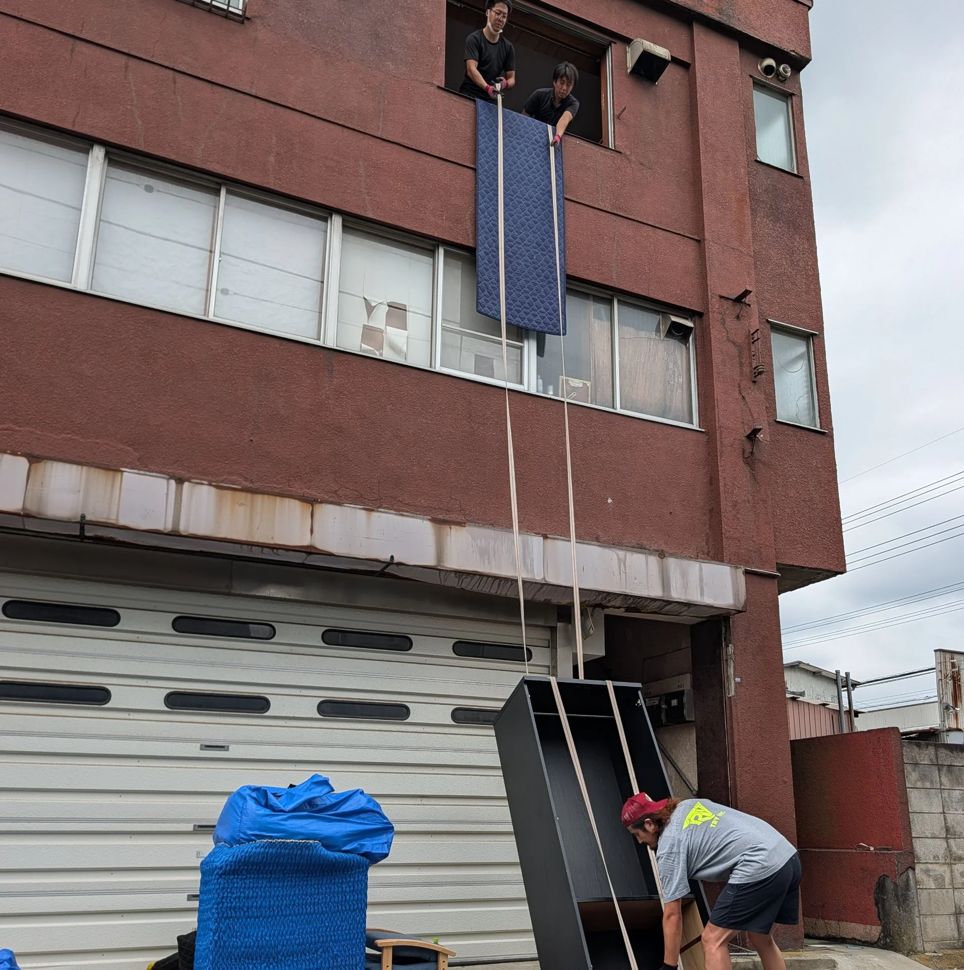 Workers lowering a large piece of furniture by rope from an upper-floor window while a ground-level worker receives it